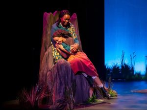 A Black woman cradles a younger Black woman in her arms on stage. The two women are wearing 1850’s period costumes and sit on a chair that looks like a tree. The lighting is low and moody. The younger person smiles, the older person appears to be crying.