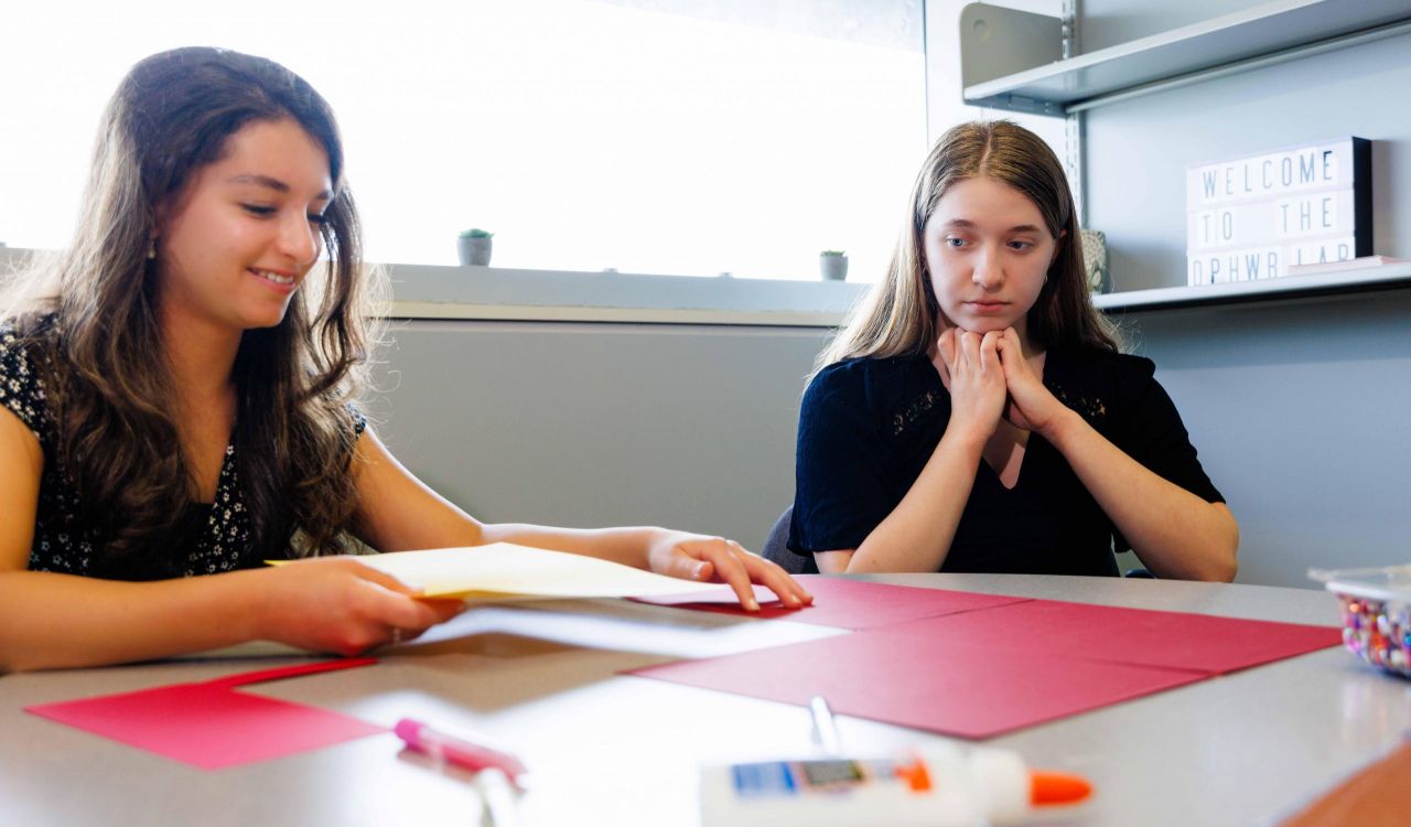 Two teenage girls sit at a table covered in paper, markers and a bottle of glue, in preparation for what looks like an arts and crafts project.