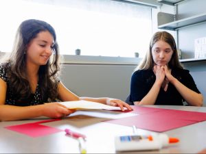 Two teenage girls sit at a table covered in paper, markers and a bottle of glue, in preparation for what looks like an arts and crafts project.