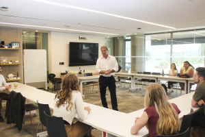 A group of university students sit at meeting tables while a man stands in the centre of the room speaking with them.