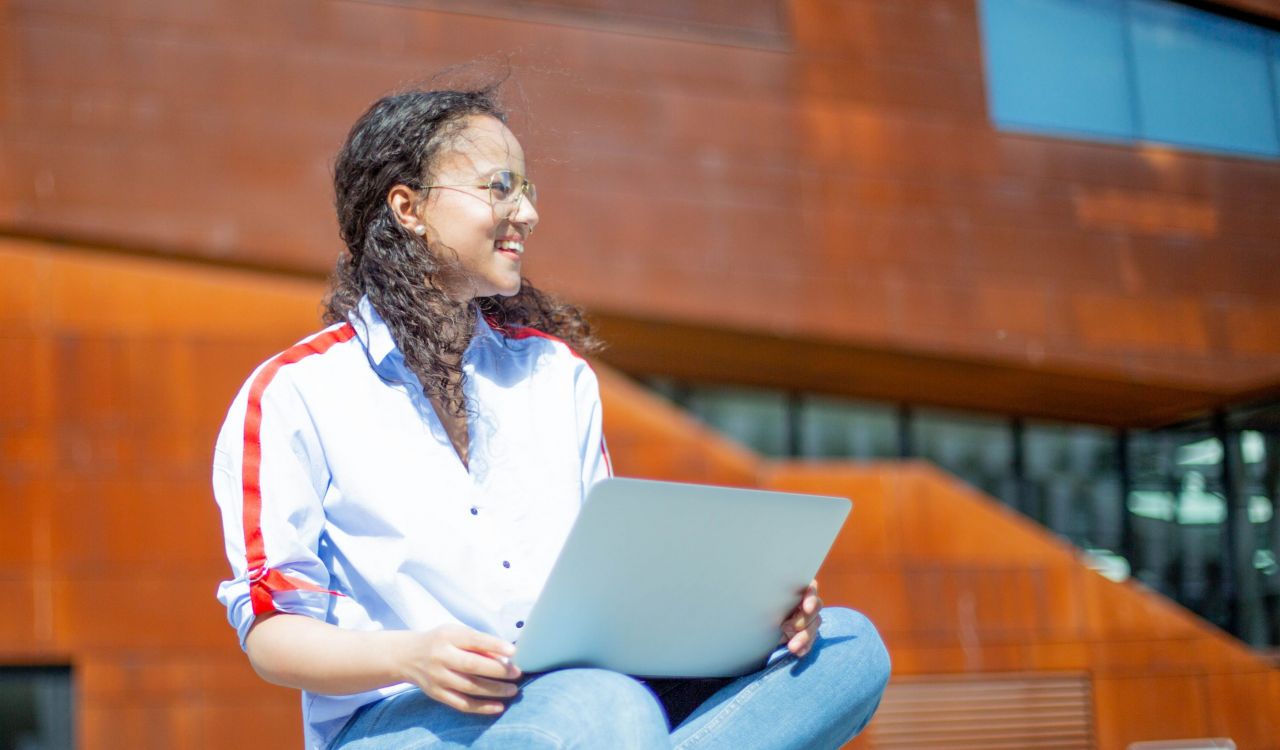 A woman sits on a bench outside with a laptop computer on her lap. The wind blows in her hair as she looks off into the distance, a smile on her face.