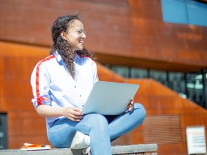 A woman sits on a bench outside with a laptop computer on her lap. The wind blows in her hair as she looks off into the distance, a smile on her face.