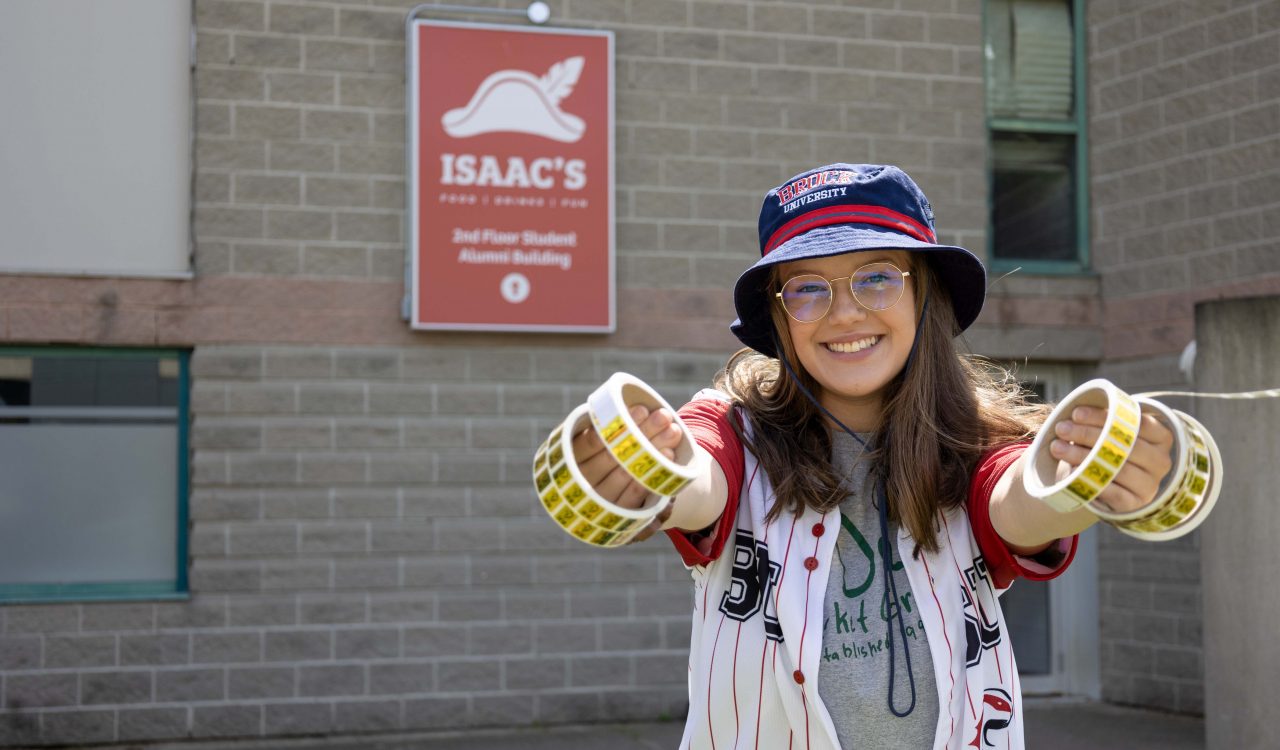 A young woman stands outside a grey building holding up rolls of gold stickers for a photo.