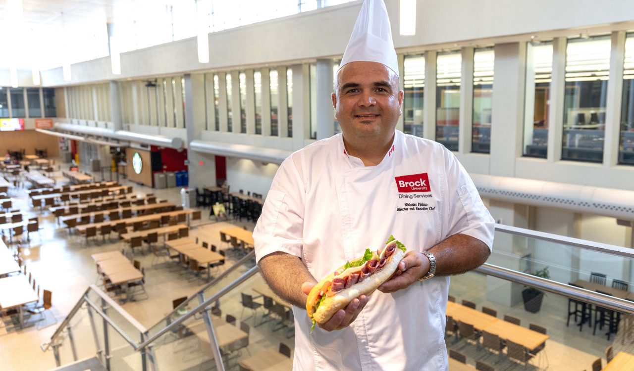 A chef holds a sub sandwich with a cafeteria behind him.