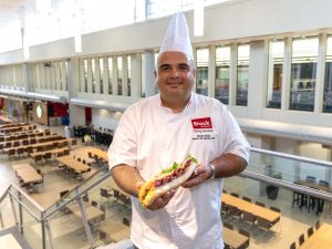 A chef holds a sub sandwich with a cafeteria behind him.