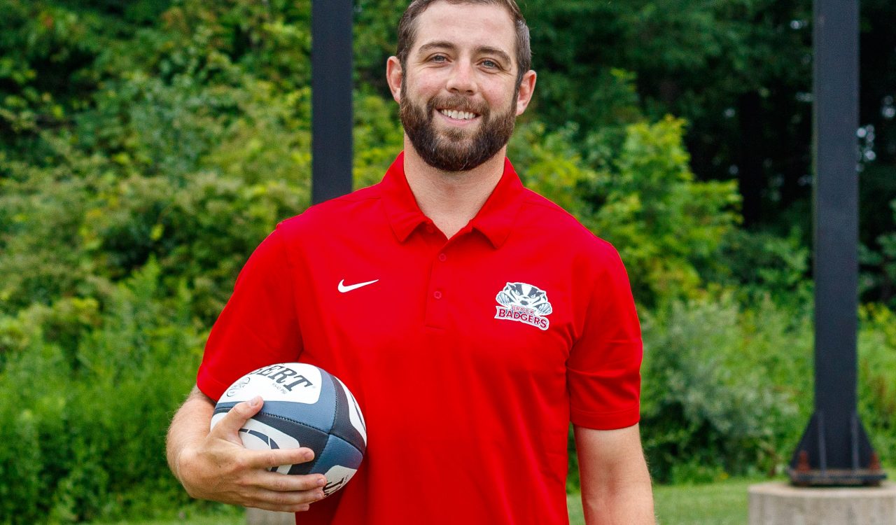 A man stands outside wearing a red polo while holding a rugby ball at Brock University.