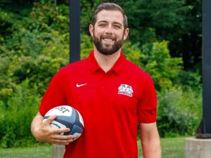 A man stands outside wearing a red polo while holding a rugby ball at Brock University.