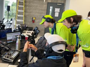 Two Brock University students volunteering at the World Rowing Championships lean over to speak with an audiovisual technician sitting in a chair and wearing a headset.