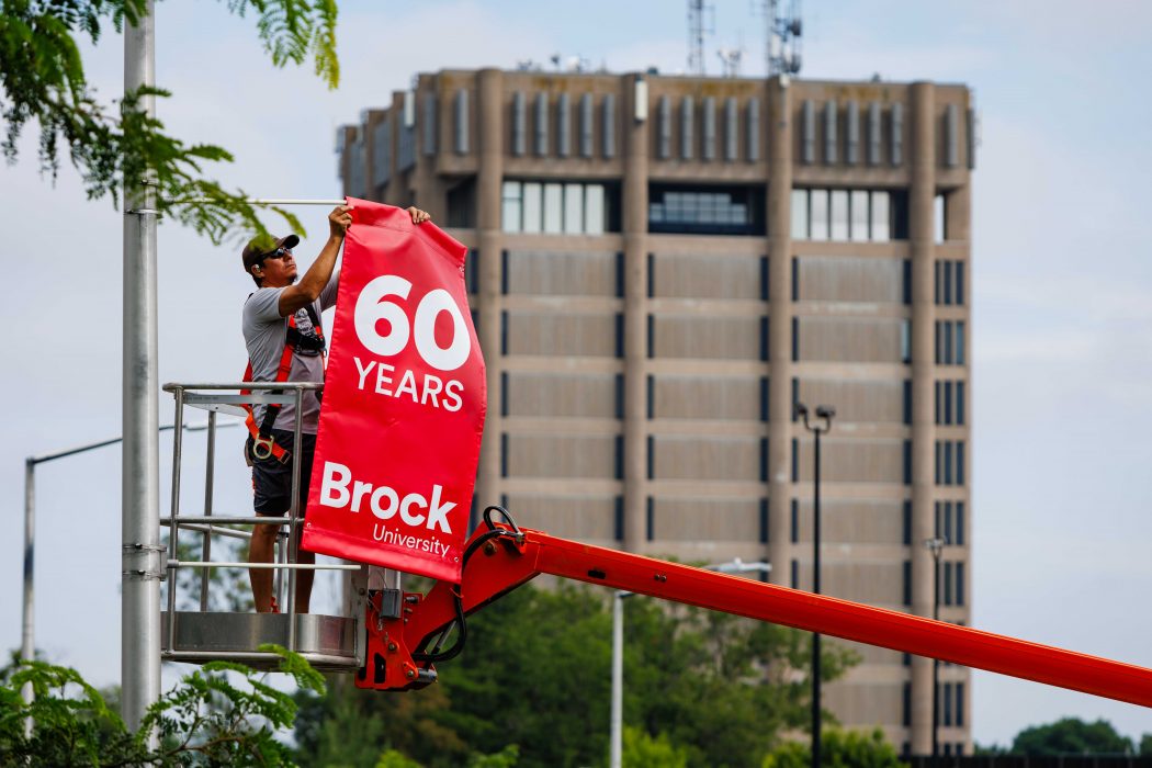 A man attaches a red banner to a light post outdoors at Brock University.