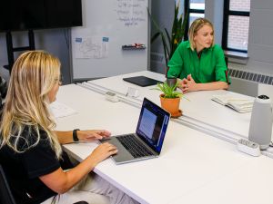 Three women sit at a table while two of them work on laptops.