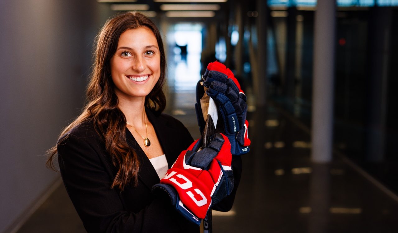 A woman stands in a glass hallway wearing business attire and hockey gloves while holding a hockey stick.