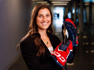 A woman stands in a glass hallway wearing business attire and hockey gloves while holding a hockey stick.