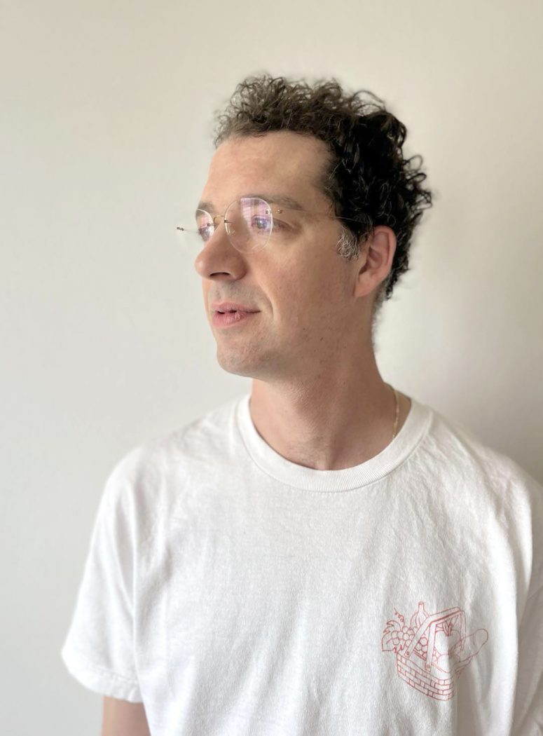 A man with curly hair wearing a plain white T-shirt. He is shown in profile, looking to the side.