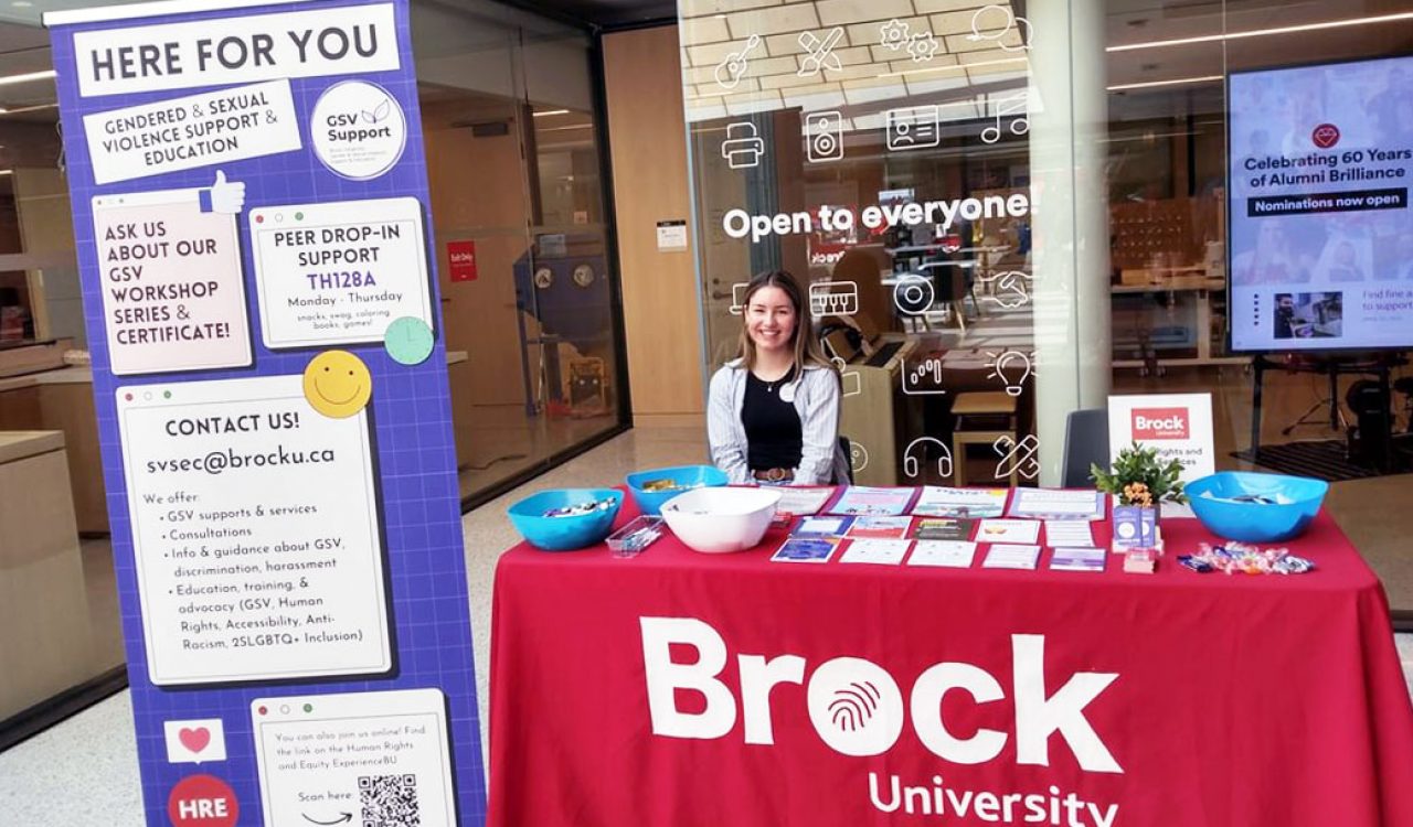 Alia Cave sits at table with tablecloth featuring a Brock logo while surrounded by posters and a pop-up banner.