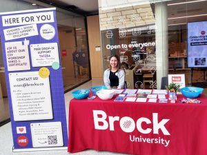 Alia Cave sits at table with tablecloth featuring a Brock logo while surrounded by posters and a pop-up banner.