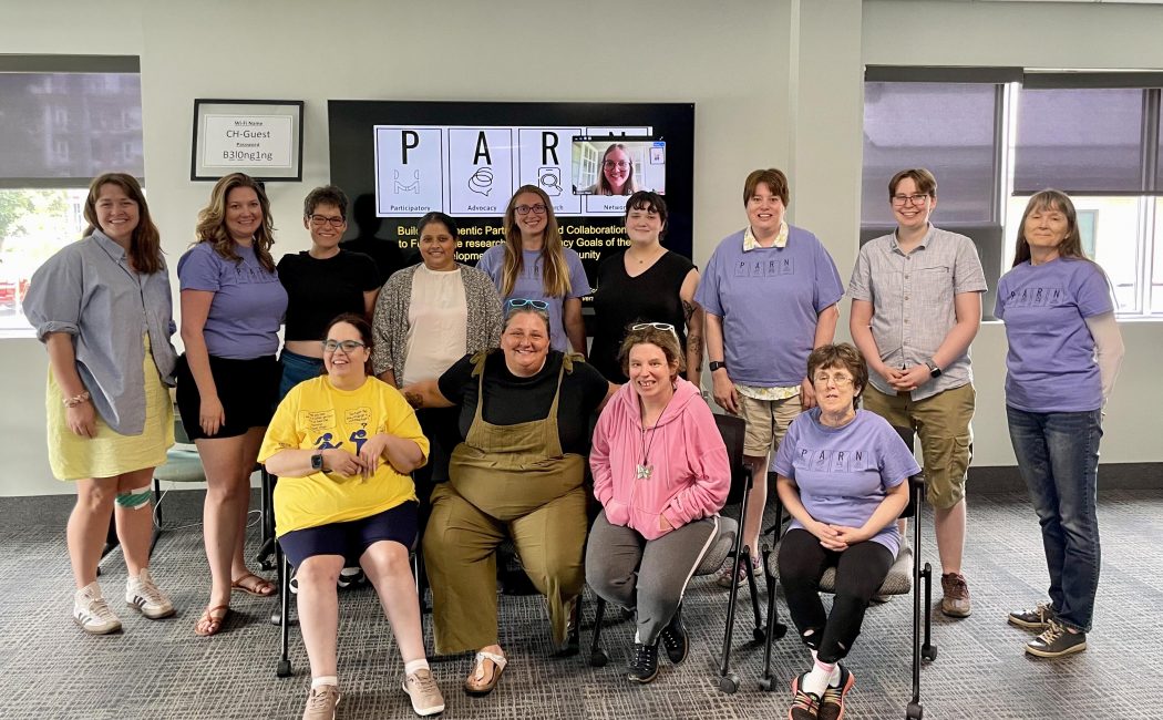 Two rows of women, one row standing in the back and one row sitting on chairs in the front, smile for the camera. Behind them is a large computer screen with the title PARN and a smaller screen showing the face of an online smiling participant. 