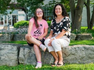 A mother and a daughter sit outside next to each other on a large, flat stone in a tree-shaded area near a university building.