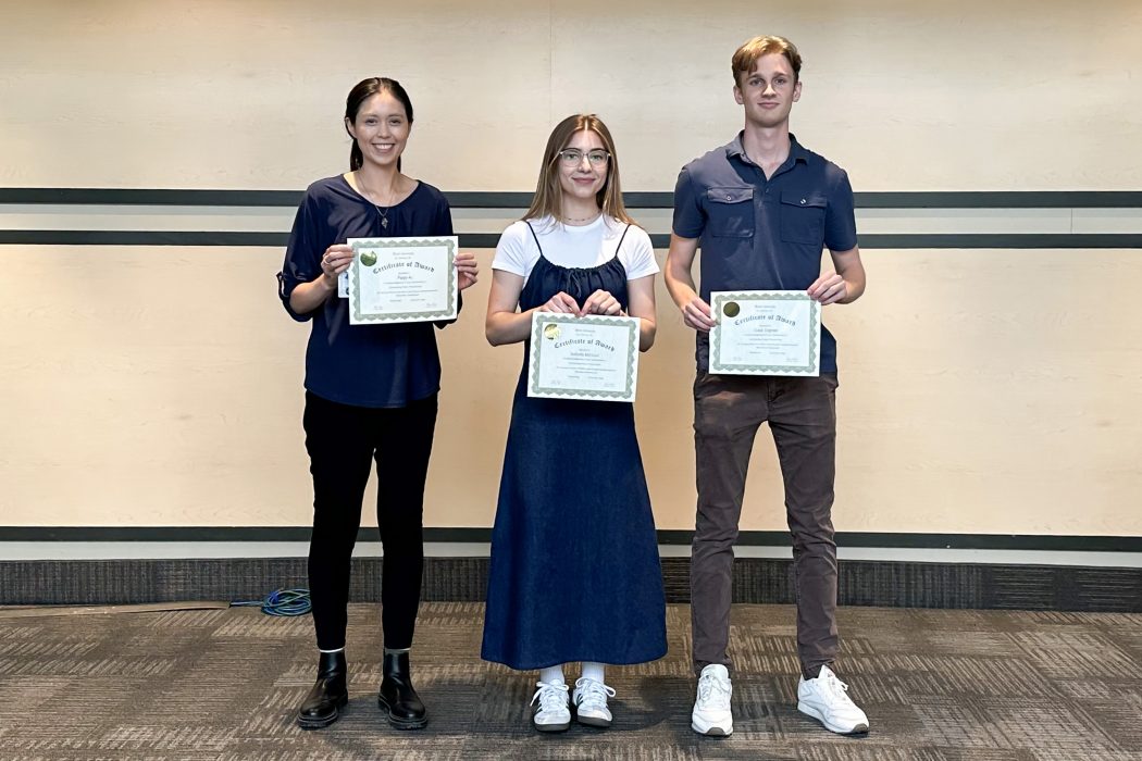 Three students stand in front of a conference room wall, each holding an award certificate.