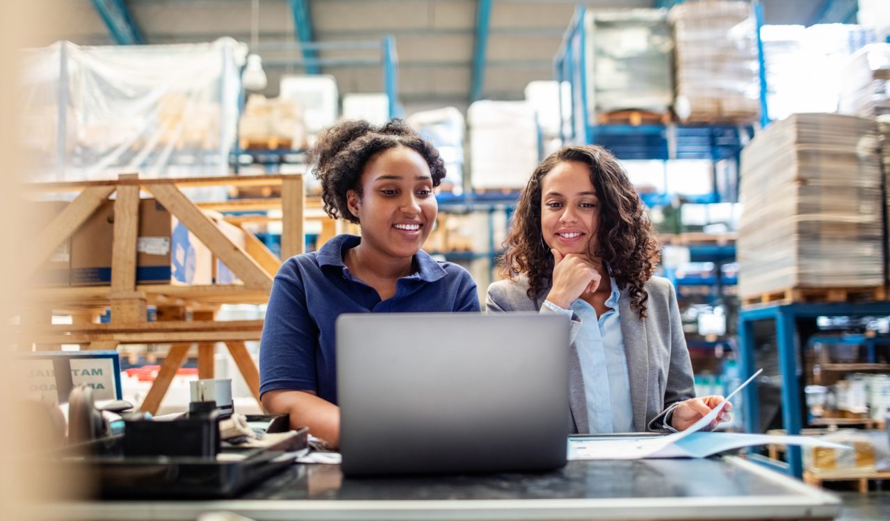 Two women work on a laptop in a warehouse.