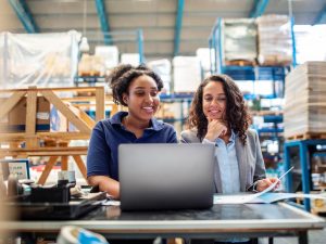 Two women work on a laptop in a warehouse.