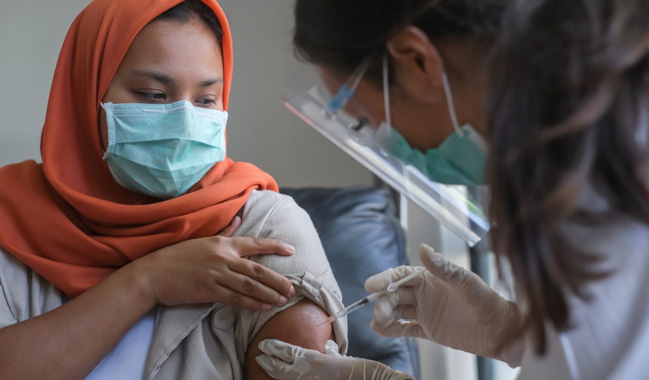 A woman in a head scarf receives a vaccine shot from a health-care worker wearing a mask and face shield.