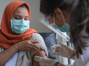 A woman in a head scarf receives a vaccine shot from a health-care worker wearing a mask and face shield.