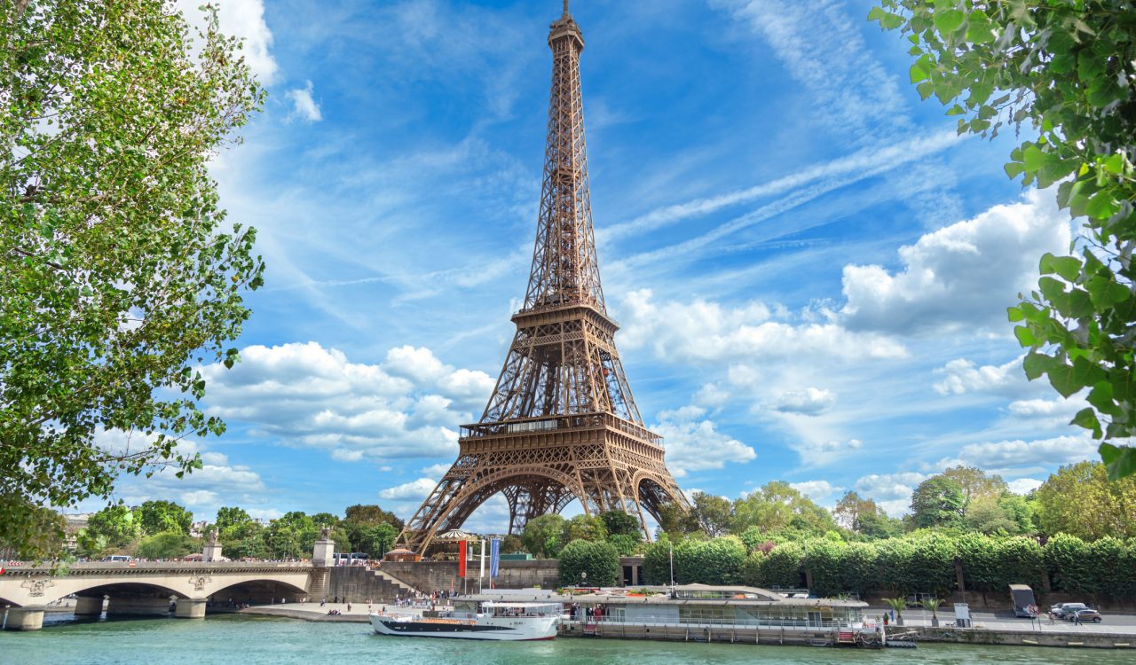 View of the Paris Eiffel Tower behind a river with boats and trees in the foreground.