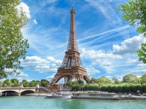 View of the Paris Eiffel Tower behind a river with boats and trees in the foreground.