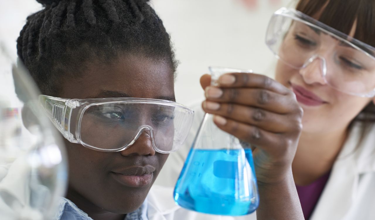 Close-up of girl’s face, wearing clear safety glasses, looking at a beaker with blue liquid she’s holding in her hand as a young woman wearing clear safety glasses looks on.