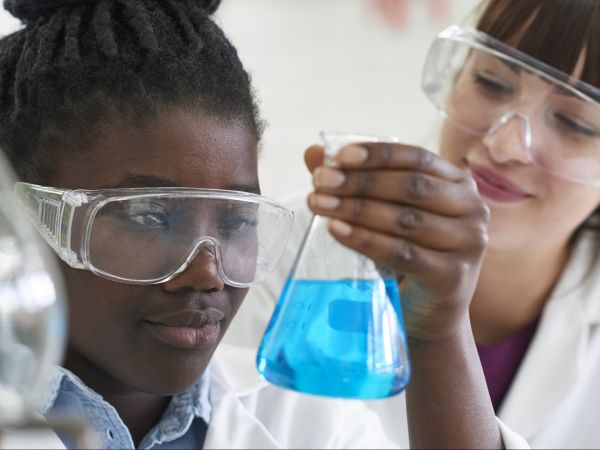 Close-up of girl’s face, wearing clear safety glasses, looking at a beaker with blue liquid she’s holding in her hand as a young woman wearing clear safety glasses looks on.