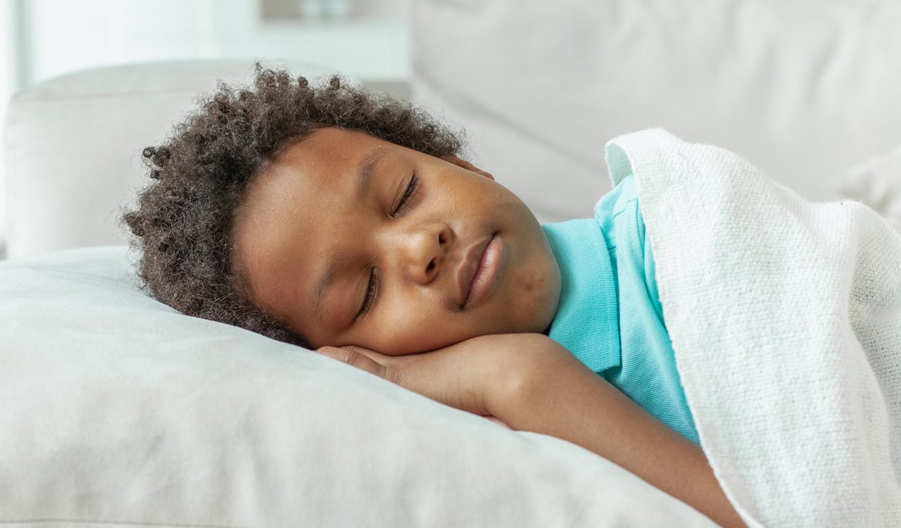 Close-up of a boy's head and shoulders resting on a pillow, with his eyes closed and his hand nestled under his face.