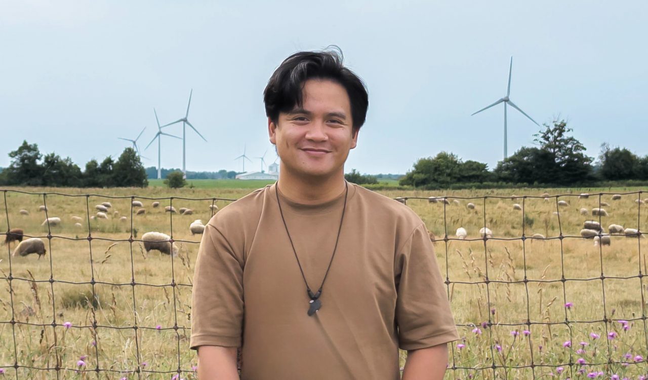 Marcus Villena stands next to a fenced farm field containing sheep. Purple wildflowers are seen in the foreground and wind turbines line the horizon in the background.