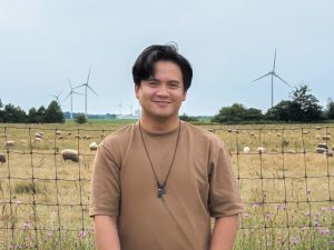 Marcus Villena stands next to a fenced farm field containing sheep. Purple wildflowers are seen in the foreground and wind turbines line the horizon in the background.