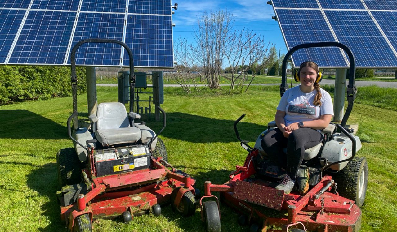 A woman sits on a riding lawnmower in front of a solar panel.