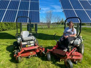 A woman sits on a riding lawnmower in front of a solar panel.