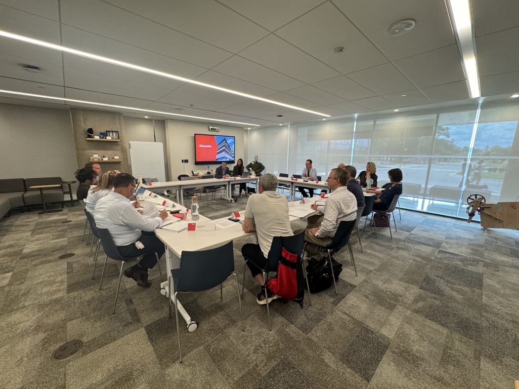 Group of people sit at a square arrangement of tables in a meeting room. A screen in the background reads "Discussion".