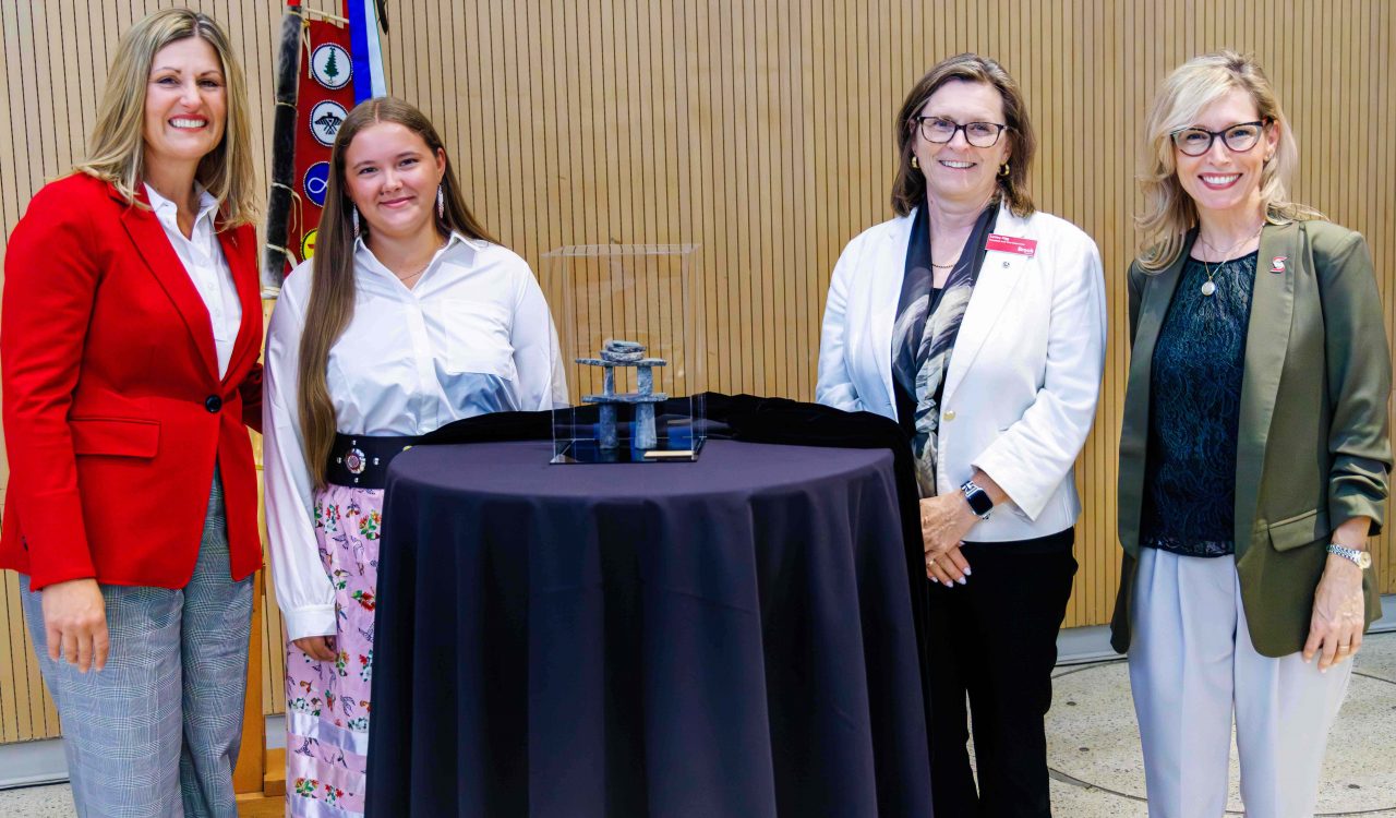 Four people stand behind a table that has an Inukshuk incased in plexiglass on it.