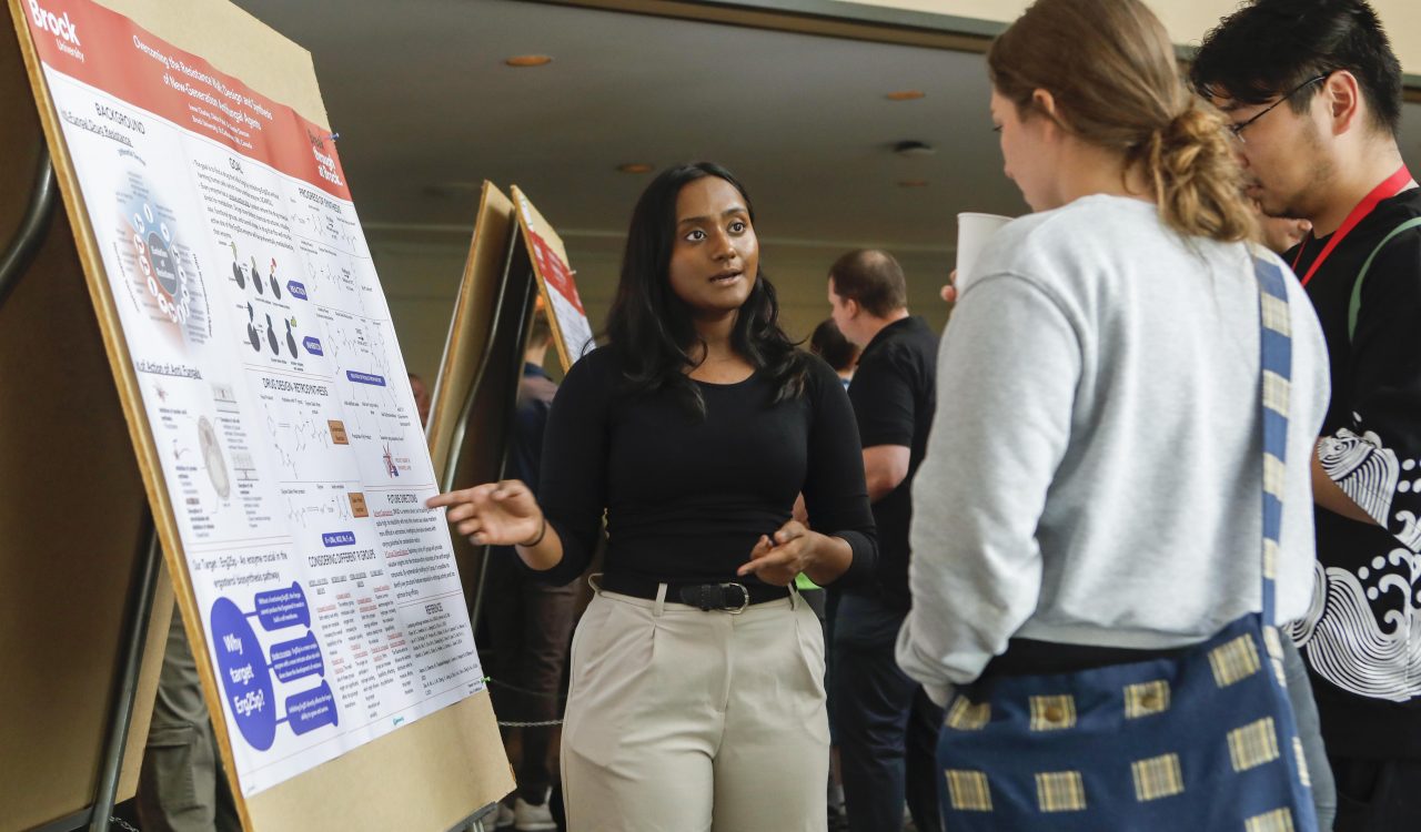 A university student points to a research poster as she speaks to people about it.