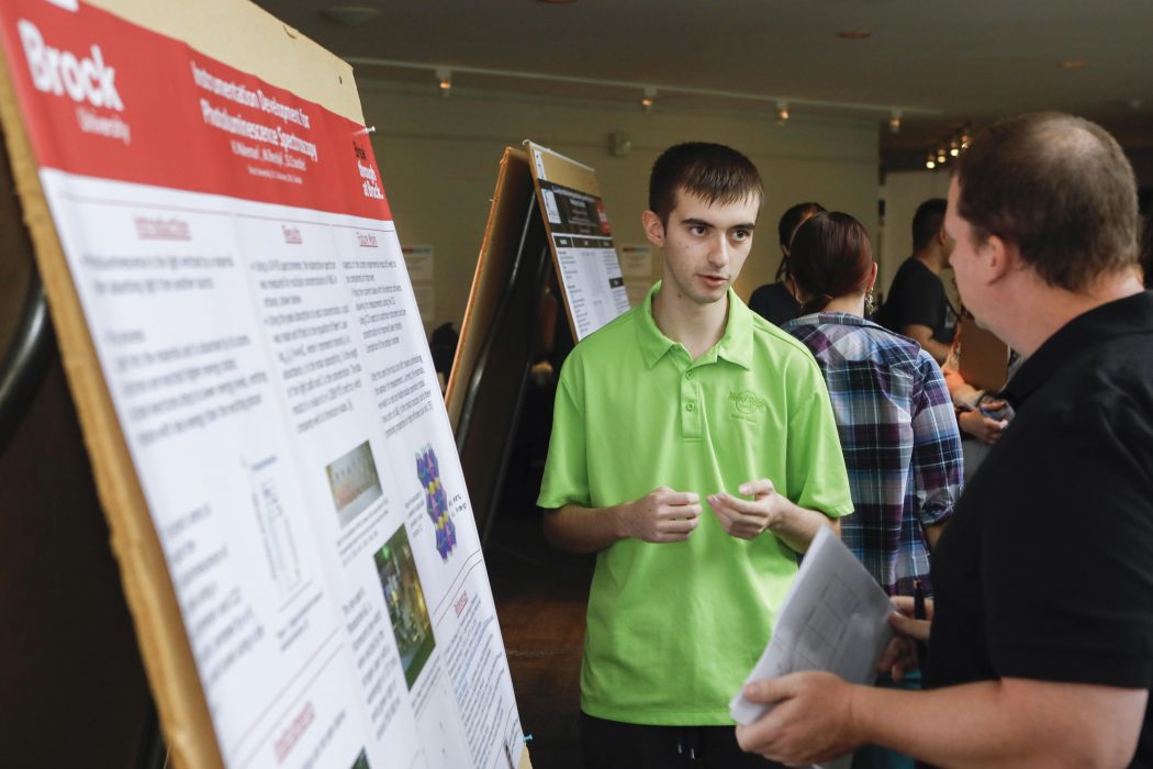 A university student points to a research poster as he speaks to people about it.