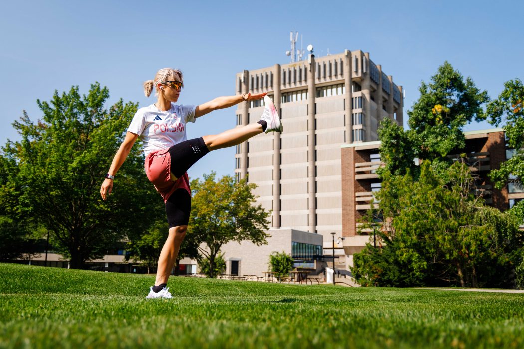 A woman stretches for a run outdoors surrounded by green trees and a blue sky on Brock University's main campus.