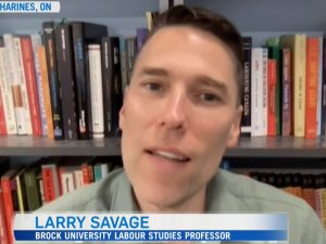 Professor Larry Savage pictured doing a television interview wearing a green collared shirt in front of a book shelf. His name and Brock University job title are shown at the bottom of the image.