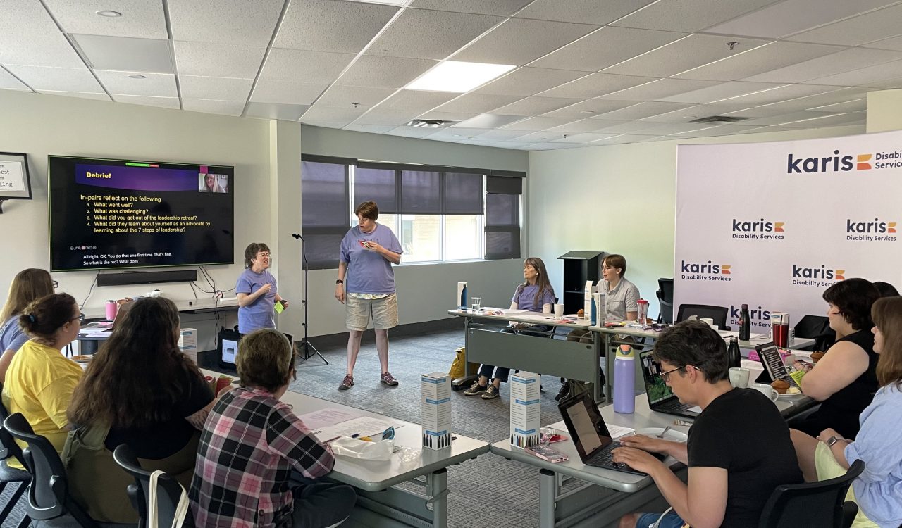 A group of people sit at tables in a meeting room with two people standing at the centre of the room.