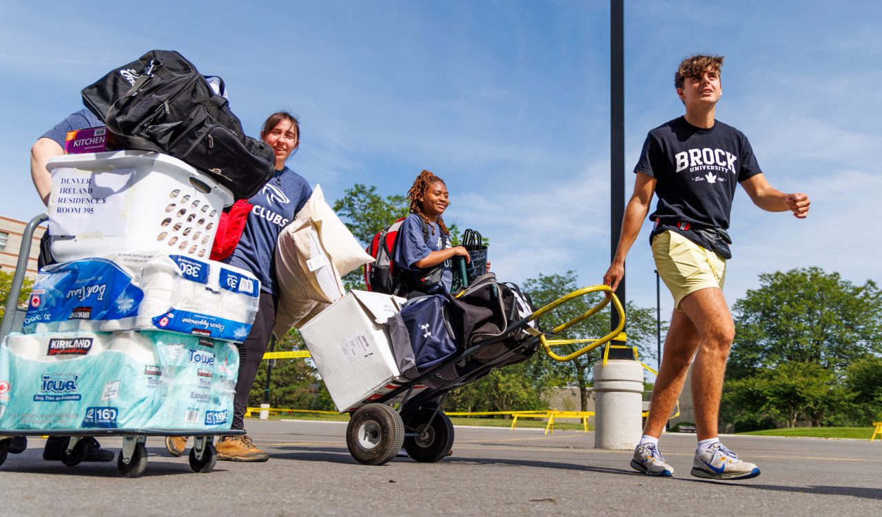 Three people pull carts loaded with bags and storage totes across a parking lot on a sunny day.