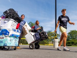 Three people pull carts loaded with bags and storage totes across a parking lot on a sunny day.