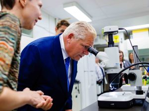 A scientist stands beside a man in a suit while he looks through a microscope.