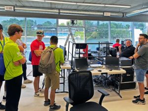 Brock University students stand in the rowing Timing Tower as they receive spectator experience training from Event 360. Desks with audiovisual equipment and computers face large windows looking out onto the Martindale Pond near the rowing competition finish line.