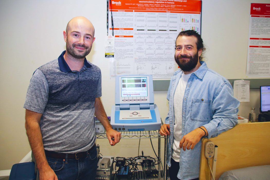 : Assistant Professor of Kinesiology Stephen Klassen (left) and his PhD student Nathan Iannarelli (right) stand in front of a wire trolly with a computer screen in the middle displaying colourful images, with a blurry research poster in the background.