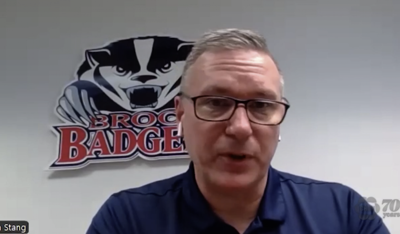 Brock fencing head coach Tim Stang wears a blue collared shirt in front of a Brock badgers logo on the wall during a video interview with CHCH news