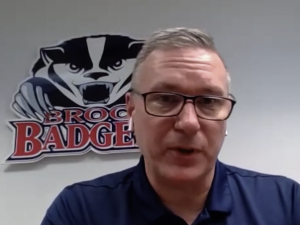 Brock fencing head coach Tim Stang wears a blue collared shirt in front of a Brock badgers logo on the wall during a video interview with CHCH news