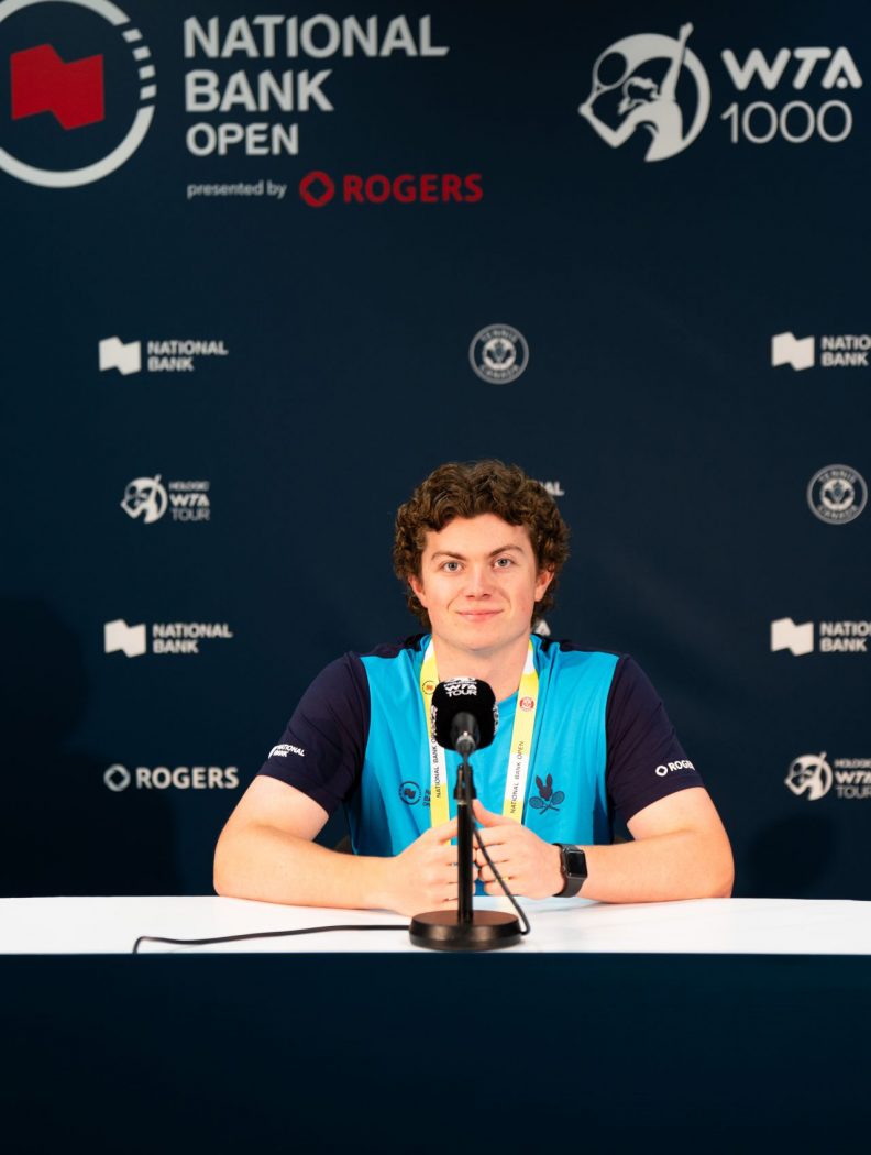 Taylor May sits at a desk with a microphone. Behind him is a backdrop that reads “National Bank Open presented by Rogers.”